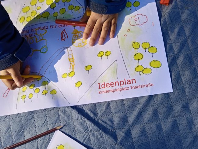 Ein Ideenplan für den Kinderspielplatz Inselstraße liegt auf einer grauen Decke, im Anschnitt malen zwei Kinderhände mit bunten Stiften eine Rutsche