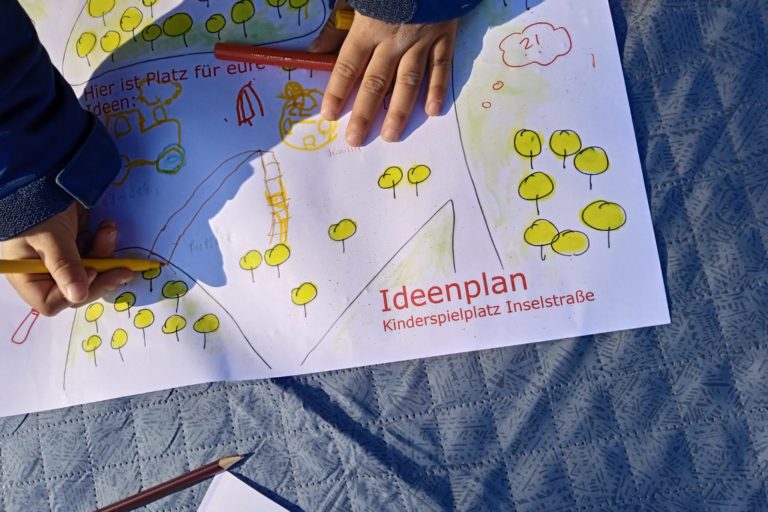 Ein Ideenplan für den Kinderspielplatz Inselstraße liegt auf einer grauen Decke, im Anschnitt malen zwei Kinderhände mit bunten Stiften eine Rutsche
