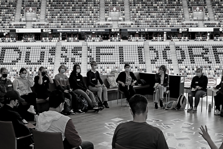 Im Vordergrund: Schulklasse im Gespräch mit Politiker:innen. Im Hintergrund: Innenraum Stadion Düsseldorf