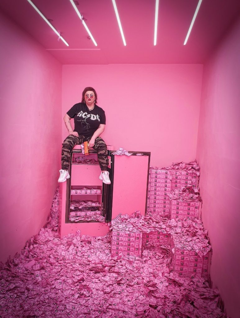 Eine Frau in einem Raum mit pinken Wänden, sitzt auf einem offenen Tresor. Um sie herum und auf dem gesamten BOden Stapeln sich Gelscheine, die auch alle pink sind.