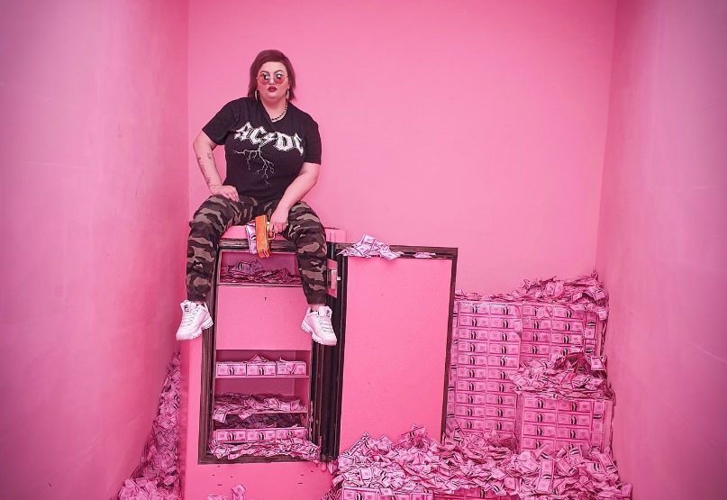Eine Frau in einem Raum mit pinken Wänden, sitzt auf einem offenen Tresor. Um sie herum und auf dem gesamten BOden Stapeln sich Gelscheine, die auch alle pink sind.