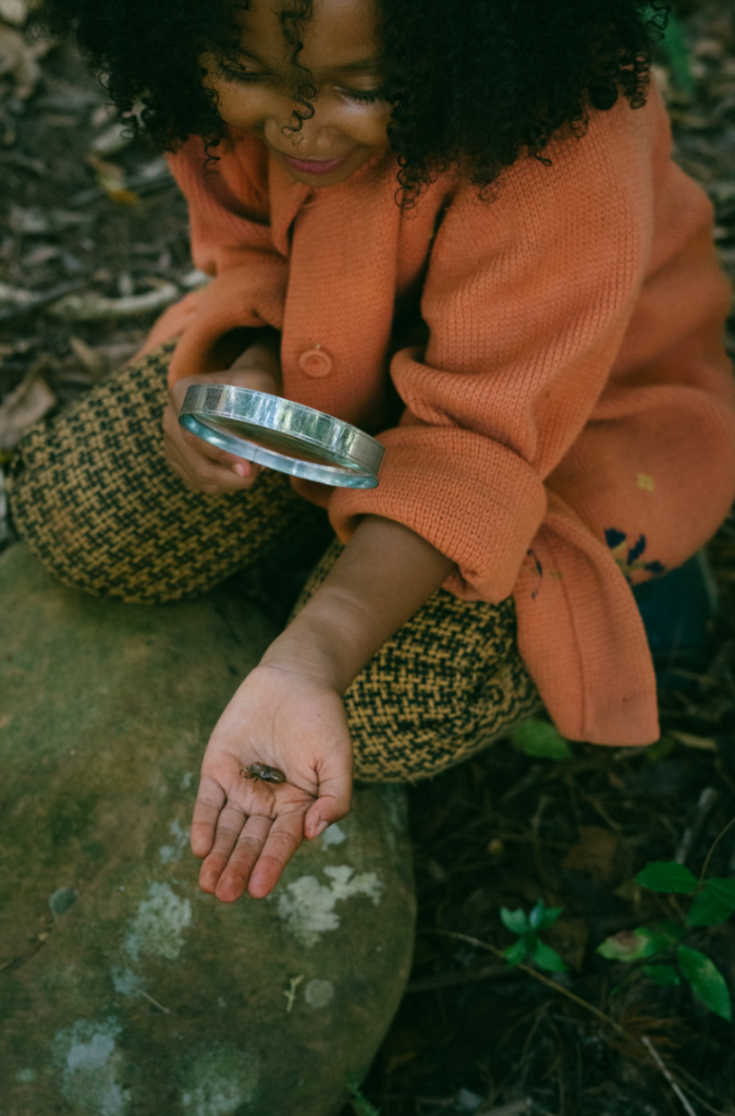 Ein Mädchen sitzt auf dem Waldboden. in ihrer linken Hand hält sie auf der Handfläche einen Käfer. In der anderen Hand hält sie eine Lupe.