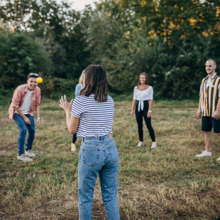 Auf einer Wiese stehen sieben junge Erwachsene in einem Kreis und spielen ein Gruppenspiel mit einem kleinen Ball