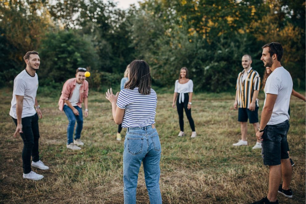 Auf einer Wiese stehen sieben junge Erwachsene in einem Kreis und spielen ein Gruppenspiel mit einem kleinen Ball