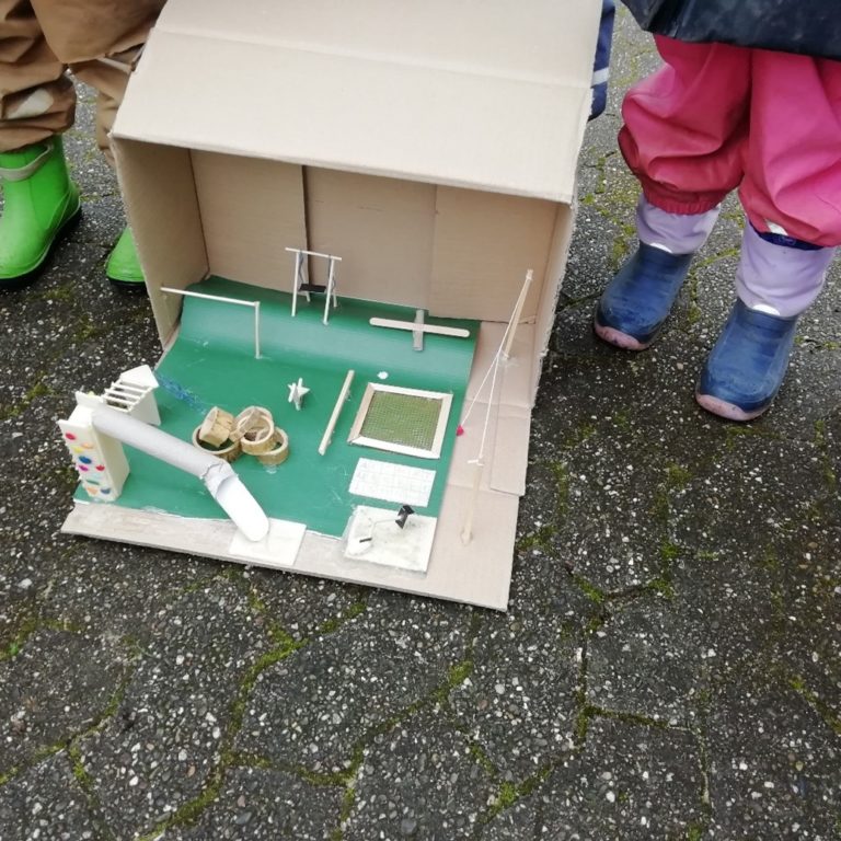 Kinder in Matschhosen und Gummistiefel stehen um ein sehr Detailgetreues Modell eines Spielplatzes herum.