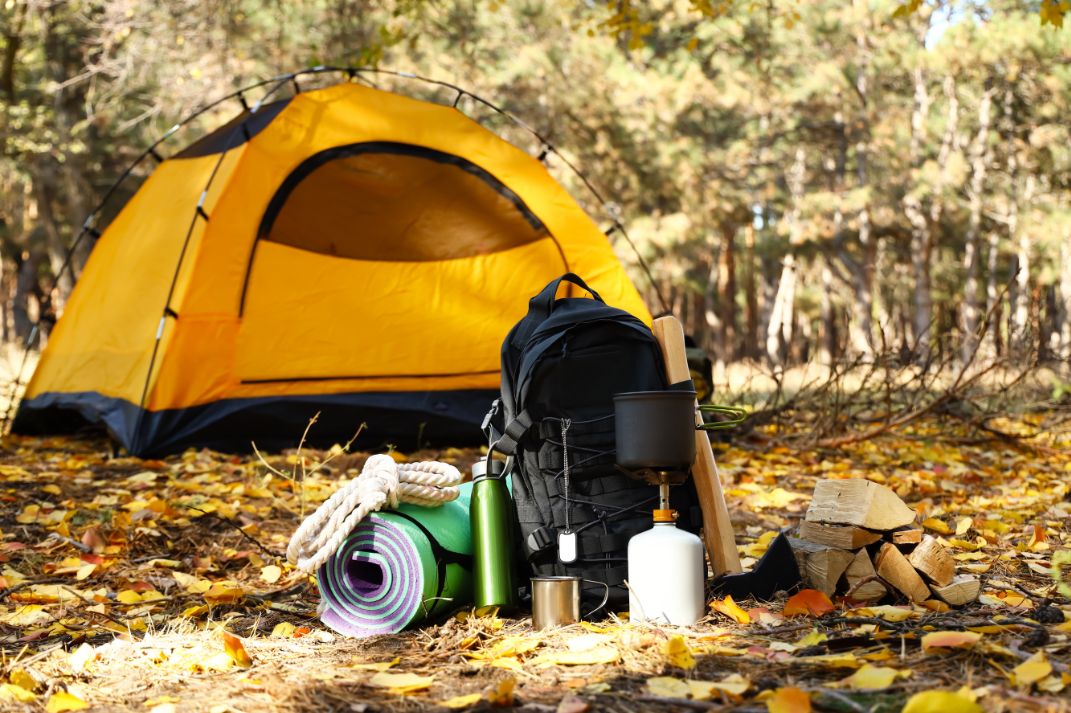 Auf einer Waldlichtung steht ein Zelt und im Vordergrund stehen eine Isomatte, Seil, Trinkflasche und ein Rucksack.