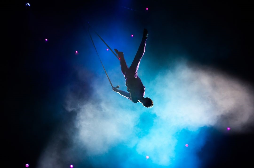 Eine Person schwingt Kopfüber an Seilen und macht Akrobatik. Der Hintergrund ist dunkel mit blauen Lichtern und liegt im Nebel.
