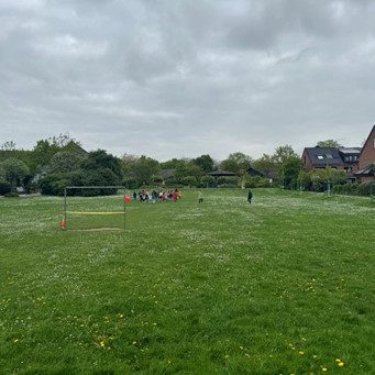 Auf der Wiese des Quartierplatzes ist in einiger Entfernung eine größere Grppe von Kindern und Erwachsenen zu sehen. Der Platz ist umgeben von Wohnhäusern und auf der Wiese steht ein Tor.