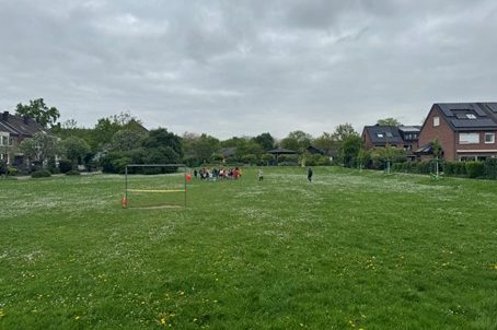 Auf der Wiese des Quartierplatzes ist in einiger Entfernung eine größere Grppe von Kindern und Erwachsenen zu sehen. Der Platz ist umgeben von Wohnhäusern und auf der Wiese steht ein Tor.