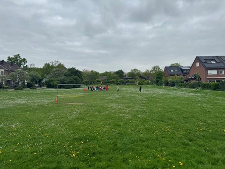 Auf der Wiese des Quartierplatzes ist in einiger Entfernung eine größere Grppe von Kindern und Erwachsenen zu sehen. Der Platz ist umgeben von Wohnhäusern und auf der Wiese steht ein Tor.